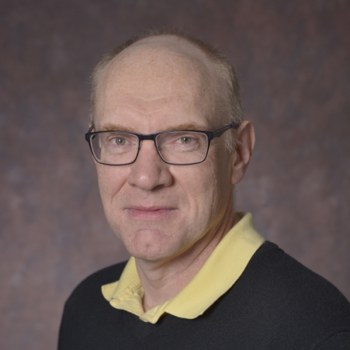 Headshot of balding male wearing eyglasses, a black pullover sweater, and a yellow shirt.