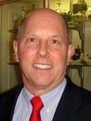 Headshot of bald male wearing a black suit jacket, light blue shirt, and a red tie.