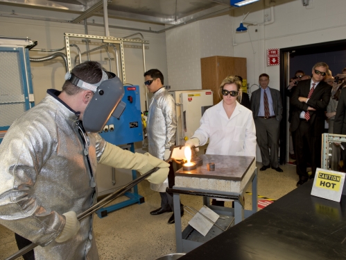 Male engineer wearing alumnized safety jacket, long-sleeved gloves, and a welding helmut as he pours molten glass from a crucible into a mold as a female engineer assists him with several onlookers observing the procedure. 