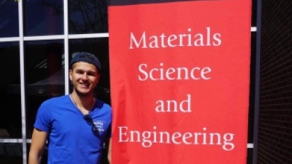 Male college student wearing a blue polo shirt, baseball cap and blue jeans stands next to a vertical banner that says Rutgers Materials Science and Engineering.