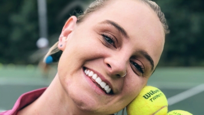 Head shot of a woman on a tennis court holding three yellow tennis balls by her head, smiling and wearing a pink top.