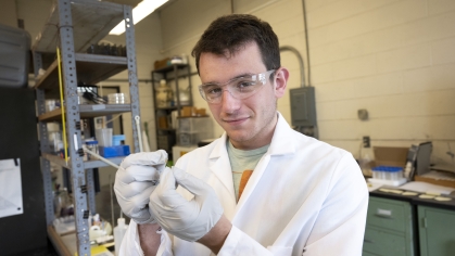 Male student working in laboratory wearing safety goggles, a white lab coat, and gloves.