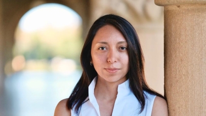 Young woman with long black hair wearing a sleeveless white blouse with her arms crossed leans against a column.