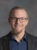 Headshot of male with facial hair and eyeglasses, wearing a dark suit and a dark bblue button down shirt.