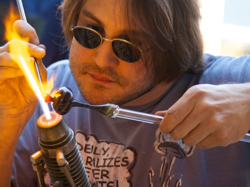 Male wearing dark safety glasses while melting a small piece of glass with high flame. 