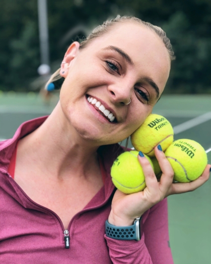 Head shot of a woman on a tennis court holding three yellow tennis balls by her head, smiling and wearing a pink top.