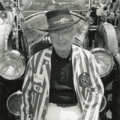 Black and white photo of elderly man wearing a stripped suit jacket and hat in the background is a vintage car.